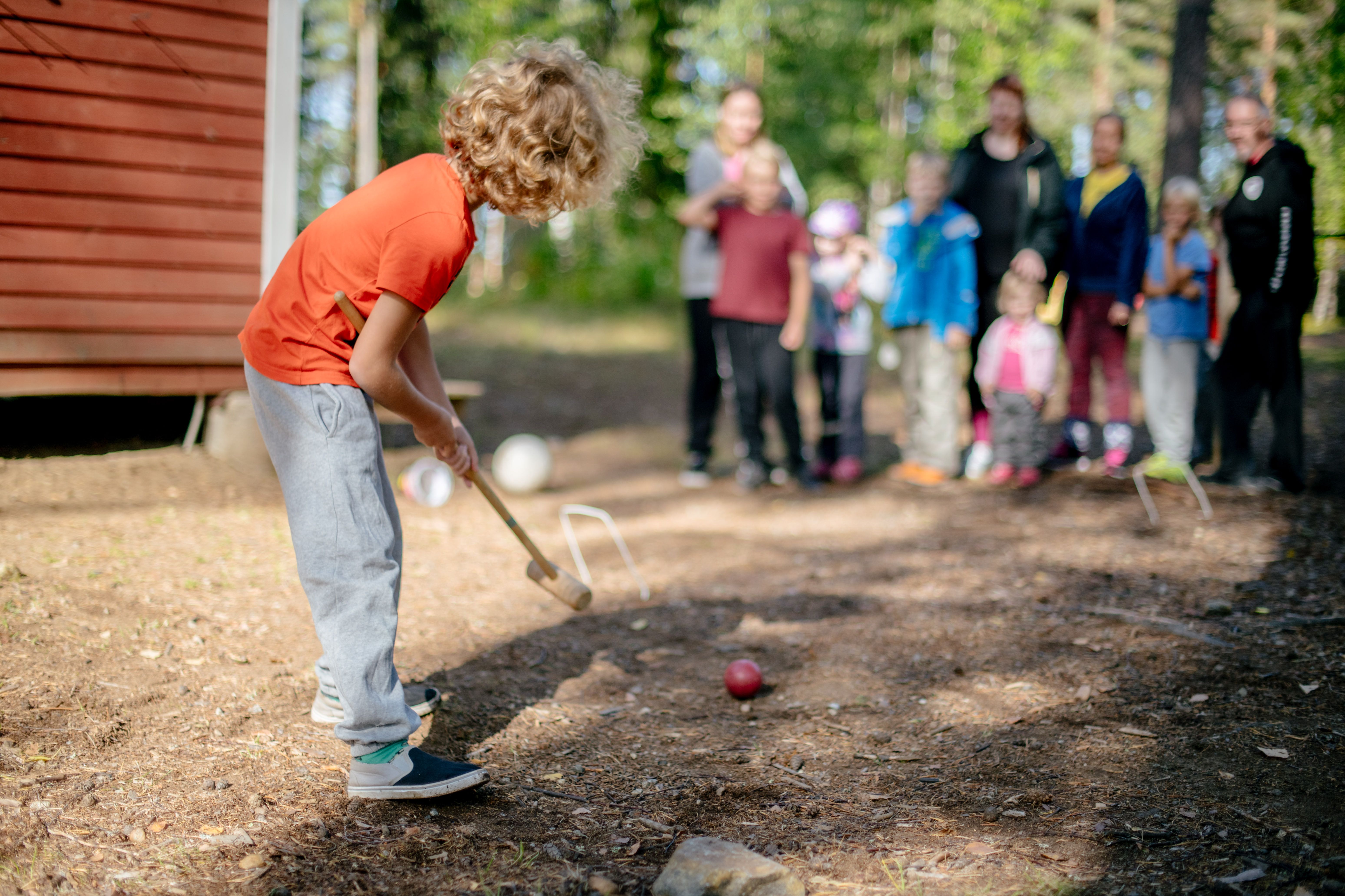 Oranssiin paitaan ja harmaisiin verkkareihin pukeutunut kiharahiuksinen lapsi pelaa krokettia kesäisessä pihapiirissä. Taustalla aikuisia ja lapsia katsoo kohti lasta.